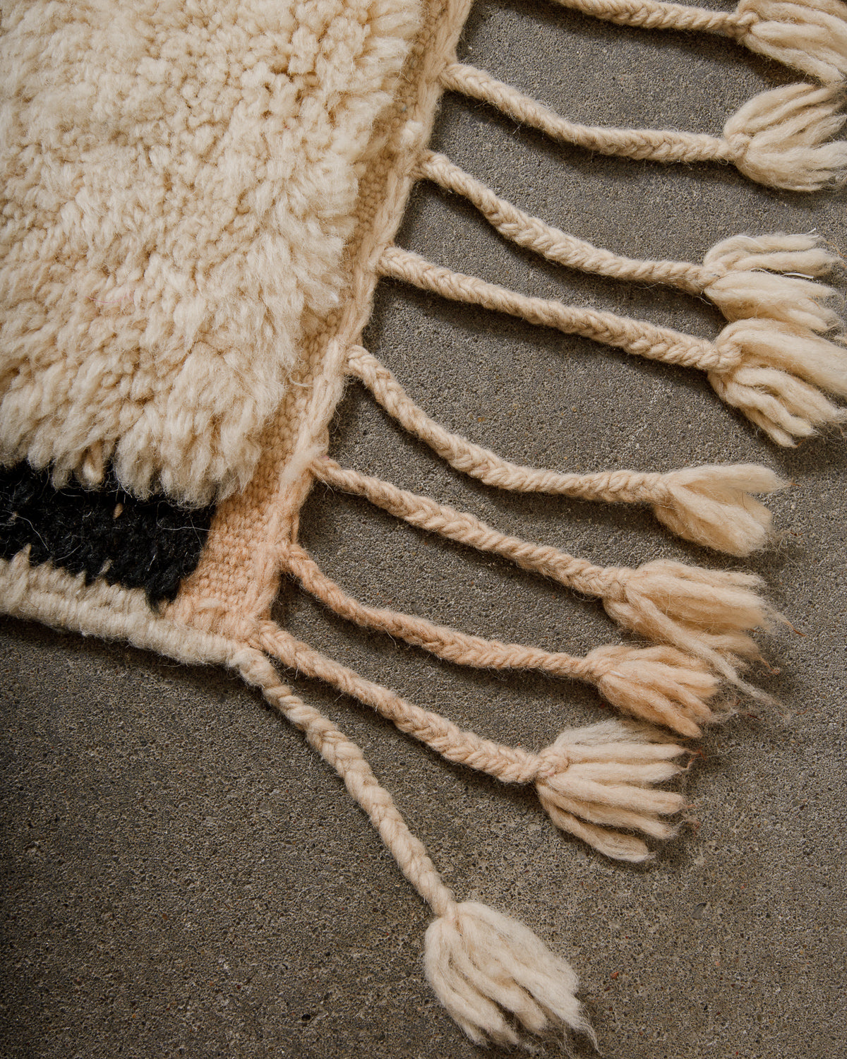 Corner of a wool rug with tassels and a black design that has discoloured part of the tassels.
